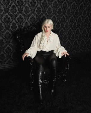 Sarışın kadın modelin klasik gotik fantezi bluzu, deri pantolonu ve beyaz sarı saçlarıyla tam boy portresi. Zarif oturma pozu, siyah taht sandalyesi. Duvar kağıdının üzerinde.