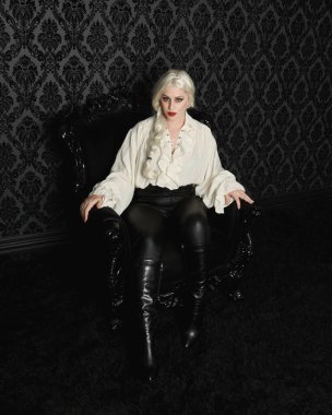 Full length portrait of blonde female model wearing vintage gothic fantasy blouse, leather pants and white blonde hair. elegant sitting pose black ornate throne chair on brocade wallpaper backdrop clipart