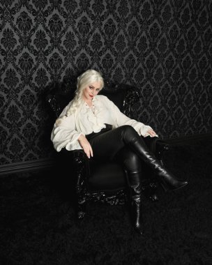 Sarışın kadın modelin klasik gotik fantezi bluzu, deri pantolonu ve beyaz sarı saçlarıyla tam boy portresi. Zarif oturma pozu, siyah taht sandalyesi. Duvar kağıdının üzerinde.
