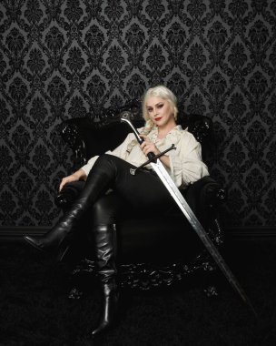  Sarışın kadın modelin klasik gotik fantezi bluz ve deri pantolon giymiş tam boy portresi. Zarif oturma pozu siyah taht koltuğu, kılıç silahı, desenli duvar kağıdı fon.