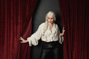 Güzel sarışın kadın modelin portresi, klasik gotik fantezi fırfırlı bluz beyaz gömlek ve deri pantolon giyiyor. Kırmızı kadife perde arkasında el hareketleriyle poz vermek.