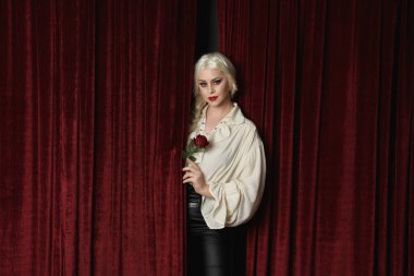 close up portrait of beautiful blonde female model, wearing vintage gothic fantasy ruffle blouse white shirt and leather pants.  Holding romantic rose, isolated against red velvet stage curtain backdrop. clipart