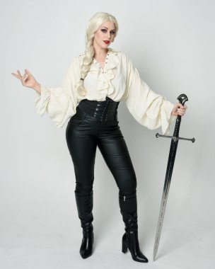 Klasik gotik fantezi bluz, deri pantolon, çizme ve sarı örgü saç giyen güzel bir kadın modelin tam boy portresi. Beyaz stüdyo arka planında izole edilmiş, savaş pozisyonunda kılıç pozisyonu almış.. 