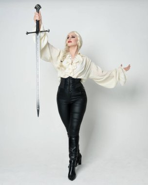 Klasik gotik fantezi bluz, deri pantolon, çizme ve sarı örgü saç giyen güzel bir kadın modelin tam boy portresi. Beyaz stüdyo arka planında izole edilmiş, savaş pozisyonunda kılıç pozisyonu almış.. 