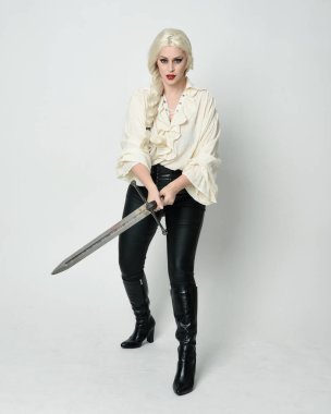 Klasik gotik fantezi bluz, deri pantolon, çizme ve sarı örgü saç giyen güzel bir kadın modelin tam boy portresi. Beyaz stüdyo arka planında izole edilmiş, savaş pozisyonunda kılıç pozisyonu almış.. 