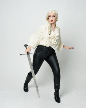 Klasik gotik fantezi bluz, deri pantolon, çizme ve sarı örgü saç giyen güzel bir kadın modelin tam boy portresi. Beyaz stüdyo arka planında izole edilmiş, savaş pozisyonunda kılıç pozisyonu almış.. 