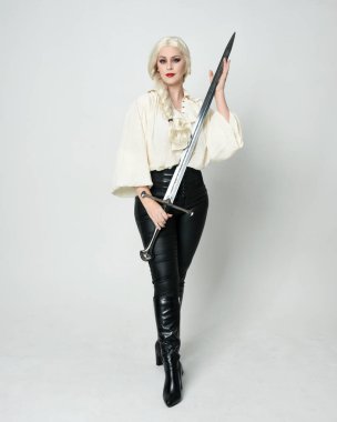 Klasik gotik fantezi bluz, deri pantolon, çizme ve sarı örgü saç giyen güzel bir kadın modelin tam boy portresi. Beyaz stüdyo arka planında izole edilmiş, savaş pozisyonunda kılıç pozisyonu almış.. 