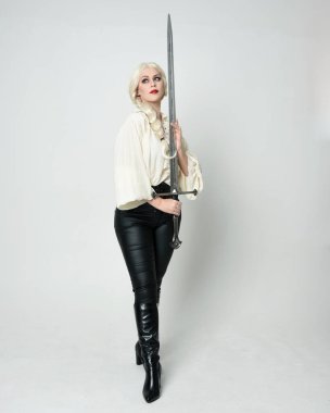 Klasik gotik fantezi bluz, deri pantolon, çizme ve sarı örgü saç giyen güzel bir kadın modelin tam boy portresi. Beyaz stüdyo arka planında izole edilmiş, savaş pozisyonunda kılıç pozisyonu almış.. 
