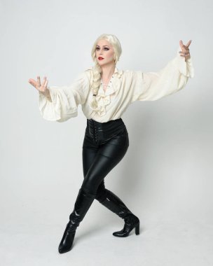  Klasik gotik fantezi bluz giyen, siyah deri pantolon giyen, sarı saçlı güzel bir kadın modelin tam boy portresi. Ayakta duran, ileri yürüyen, beyaz stüdyo arka planında izole edilmiş..