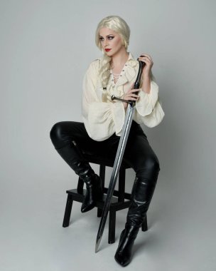 Klasik gotik fantezi bluz, siyah deri pantolon ve beyaz sarı saçlı kadın modelin tam boy portresi. Oturma pozisyonu kılıçlı silah, aksiyon pozu, stüdyo arka planında izole..