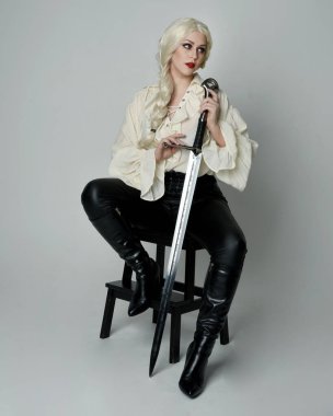 Klasik gotik fantezi bluz, siyah deri pantolon ve beyaz sarı saçlı kadın modelin tam boy portresi. Oturma pozisyonu kılıçlı silah, aksiyon pozu, stüdyo arka planında izole..