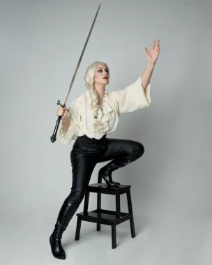 Klasik gotik fantezi bluz, siyah deri pantolon ve beyaz sarı saçlı kadın modelin tam boy portresi. Oturma pozisyonu kılıçlı silah, aksiyon pozu, stüdyo arka planında izole..