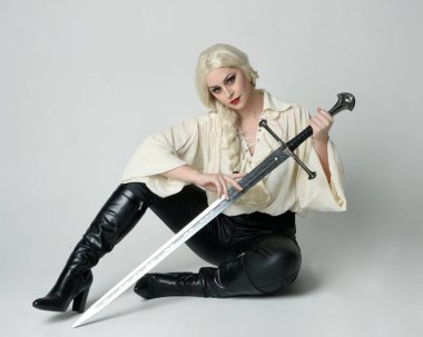 Klasik gotik fantezi bluz, deri pantolon ve beyaz sarı saç giyen, diz çökmüş kılıç tutan, stüdyo arka planında izole edilmiş kadın modelin tam boy portresi..