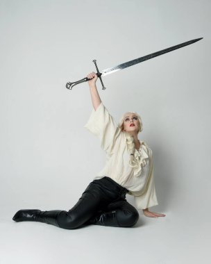 Klasik gotik fantezi bluz, deri pantolon ve beyaz sarı saç giyen, diz çökmüş kılıç tutan, stüdyo arka planında izole edilmiş kadın modelin tam boy portresi..