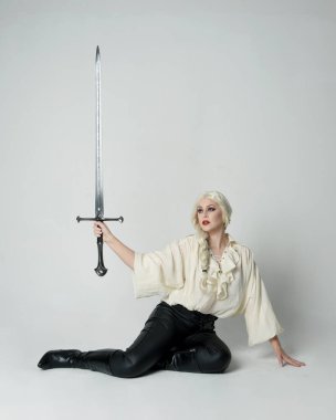 Klasik gotik fantezi bluz, deri pantolon ve beyaz sarı saç giyen, diz çökmüş kılıç tutan, stüdyo arka planında izole edilmiş kadın modelin tam boy portresi..