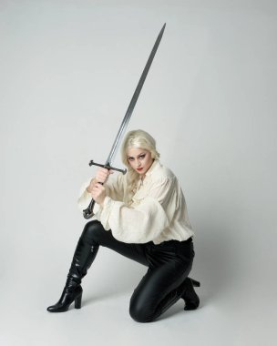 Klasik gotik fantezi bluz, deri pantolon ve beyaz sarı saç giyen, diz çökmüş kılıç tutan, stüdyo arka planında izole edilmiş kadın modelin tam boy portresi..