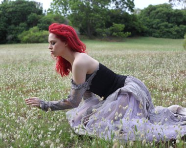 Güzel kızıl saçlı kadın modelin portresi, gotik fantezi kostümlü yırtık balo elbisesi, siyah korse. Rüya gibi çiçek tarlası orman manzarası arka planında oturuyor..