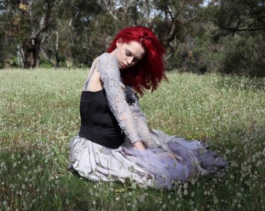 Güzel kızıl saçlı kadın modelin portresi, gotik fantezi kostümlü yırtık balo elbisesi, siyah korse. Rüya gibi çiçek tarlası orman manzarası arka planında oturuyor..