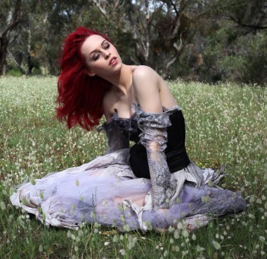 Güzel kızıl saçlı kadın modelin tam boy portresi gotik fantezi kostümlü yırtık balo elbisesi, siyah korse, deri botlar. oturma pozu orman manzarası arka planı.