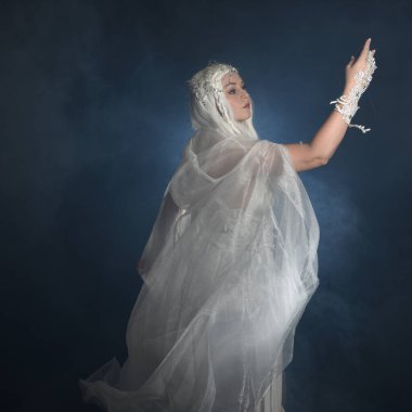 Beyaz fantezi kar perisi elbisesi giyen güzel sarışın kadın modelin portresi, sihirli gümüş kristal kraliçe tacı, kapüşonlu, katıksız pelerin. Karanlık stüdyo arka planında izole bir poz.