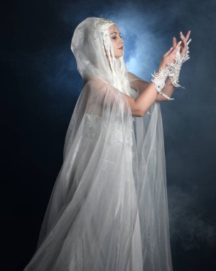 Beyaz fantezi kar perisi elbisesi giyen güzel sarışın kadın modelin portresi, sihirli gümüş kristal kraliçe tacı, kapüşonlu, katıksız pelerin. Karanlık stüdyo arka planında izole bir poz.