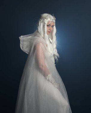 close up portrait of beautiful blonde female model wearing  white fantasy snow fairy gown,  magical crystal queen crown, hooded cloak.  Isolated pose, facing away looking back,  dark studio background clipart