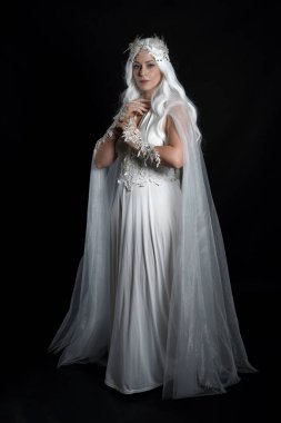 full length portrait of beautiful blonde female model wearing  white fantasy snow fairy gown,  magical silver crystal queen crown, hooded flowing cloak. Isolated walking pose dark studio background clipart