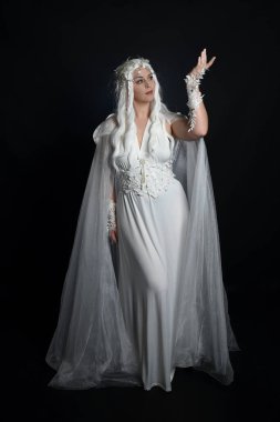 Beyaz fantezi kar perisi elbisesi, sihirli gümüş kristal kraliçe tacı ve kapüşonlu pelerin giyen güzel sarışın kadın modelin tam boy portresi. İzole edilmiş yürüyüş, karanlık stüdyo arka planı.