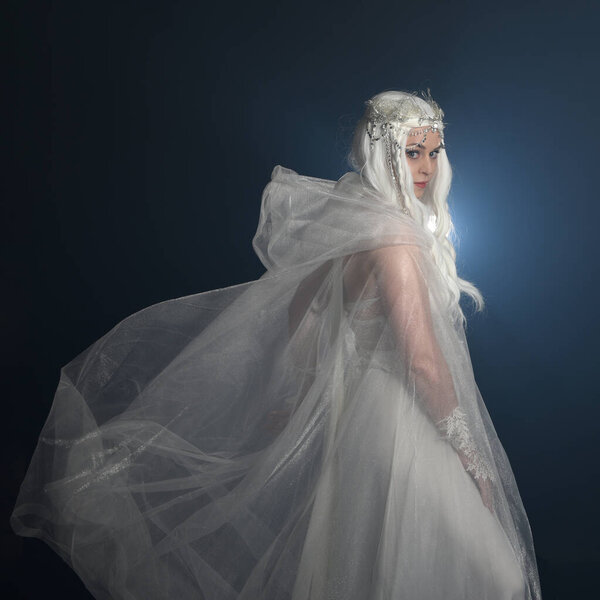 крупным планом портрет красивой блондинки-модели в белом фэнтези снежной феи, волшебная хрустальная корона королевы, плащ в капюшоне. Изолированная поза, оглядываясь назад, темный студийный фон