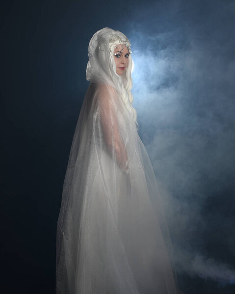 close up portrait of beautiful blonde female model wearing  white fantasy snow fairy gown,  magical crystal queen crown, hooded cloak.  Isolated pose, facing away looking back,  dark studio background