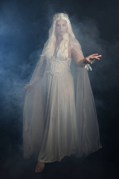 full length portrait of beautiful blonde female model wearing  white fantasy snow fairy gown,  magical silver crystal queen crown, hooded flowing cloak. Isolated walking pose dark studio background