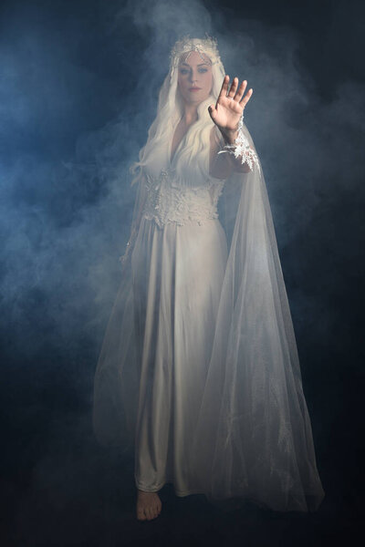 full length portrait of beautiful blonde female model wearing  white fantasy snow fairy gown,  magical silver crystal queen crown, hooded flowing cloak. Isolated walking pose dark studio background