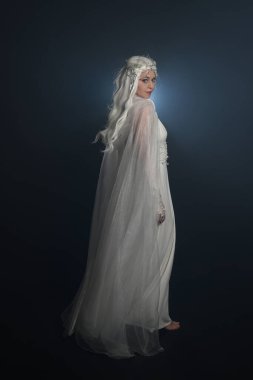 Beyaz fantezi kar perisi elbisesi giyen güzel sarışın kadın modelin tam boy portresi, sihirli gümüş kristal kraliçe tacı, kapüşonlu pelerin. İzole edilmiş yürüyüş pozisyonu, geriye dönüp bakmak, karanlık stüdyo arka planında.
