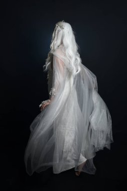 Beyaz fantezi kar perisi elbisesi giyen güzel sarışın kadın modelin tam boy portresi, sihirli gümüş kristal kraliçe tacı, kapüşonlu pelerin. İzole edilmiş yürüyüş pozisyonu, geriye dönüp bakmak, karanlık stüdyo arka planında.