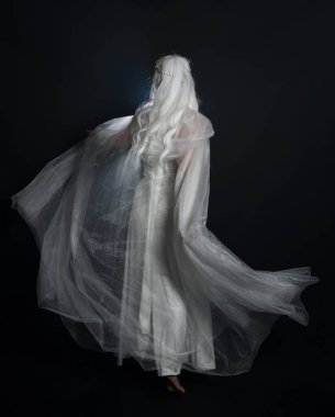 full length portrait of beautiful blonde female model wearing  white fantasy snow fairy gown,  magical silver crystal queen crown, hooded cloak. Isolated walking  pose, facing away looking back, on dark studio background. clipart