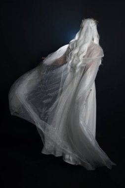 Beyaz fantezi kar perisi elbisesi giyen güzel sarışın kadın modelin tam boy portresi, sihirli gümüş kristal kraliçe tacı, kapüşonlu pelerin. İzole edilmiş yürüyüş pozisyonu, geriye dönüp bakmak, karanlık stüdyo arka planında.