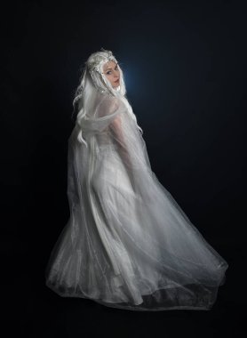 full length portrait of beautiful blonde female model wearing  white fantasy snow fairy gown,  magical silver crystal queen crown, hooded cloak. Isolated walking  pose, facing away looking back, on dark studio background. clipart