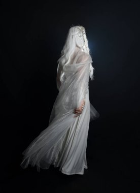 Beyaz fantezi kar perisi elbisesi giyen güzel sarışın kadın modelin tam boy portresi, sihirli gümüş kristal kraliçe tacı, kapüşonlu pelerin. İzole edilmiş yürüyüş pozisyonu, geriye dönüp bakmak, karanlık stüdyo arka planında.