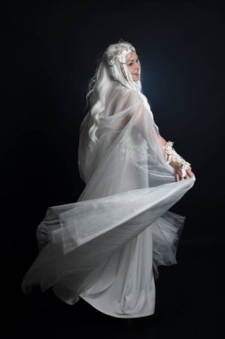 Beyaz fantezi kar perisi elbisesi giyen güzel sarışın kadın modelin tam boy portresi, sihirli gümüş kristal kraliçe tacı, kapüşonlu pelerin. İzole edilmiş yürüyüş pozisyonu, geriye dönüp bakmak, karanlık stüdyo arka planında.