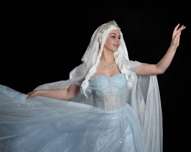 Kapalı portre güzel sarışın fantezi perisi ortaçağ peri masalı balo elbisesi giymiş, peçeli beyaz taçlı bir pelerin. Karanlık stüdyo arkaplanına uzanan solgun bir jest.