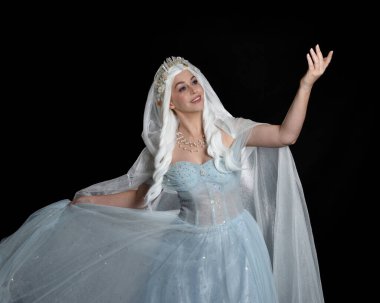 Kapalı portre güzel sarışın fantezi perisi ortaçağ peri masalı balo elbisesi giymiş, peçeli beyaz taçlı bir pelerin. Karanlık stüdyo arkaplanına uzanan solgun bir jest.