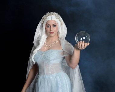Ortaçağ masal elbisesi, beyaz pelerin ve kristal taç giyen güzel sarışın fantezi perisi portresi. Işıldayan kristal küre tutarak izole karanlık stüdyo arka planı ile mistik sis arka ışık efekti