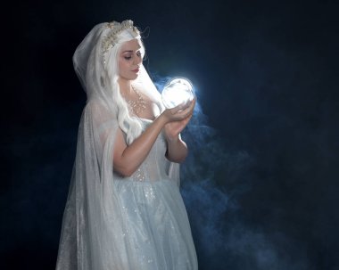 Ortaçağ masal elbisesi, beyaz pelerin ve kristal taç giyen güzel sarışın fantezi perisi portresi. Işıldayan kristal küre tutarak izole karanlık stüdyo arka planı ile mistik sis arka ışık efekti