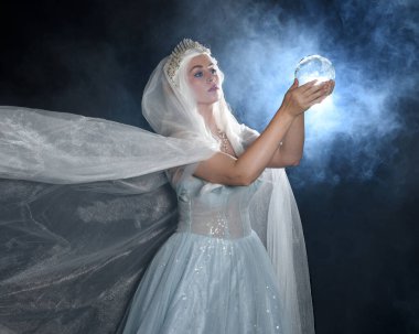 Ortaçağ masal elbisesi, beyaz pelerin ve kristal taç giyen güzel sarışın fantezi perisi portresi. Işıldayan kristal küre tutarak izole karanlık stüdyo arka planı ile mistik sis arka ışık efekti