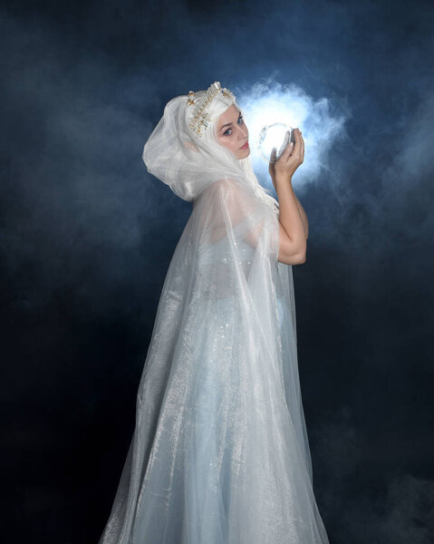 портрет красивая блондинка фантазии фея в средневековой сказке бальный костюм, белый плащ и хрустальная корона. держа светящийся хрустальный шар шар изолированный темный фон студии с мистическим дымовым туманом подсветки эффекта освещения