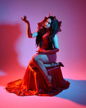 Gotik fantezi burlesk cadılar bayramı temalı şeytan kostümlü kırmızı elbise giyen güzel bir kadın manken portresi, boynuz ve eldiven. izole oturma pozisyonu, dramatik renkli stüdyo ışıkları