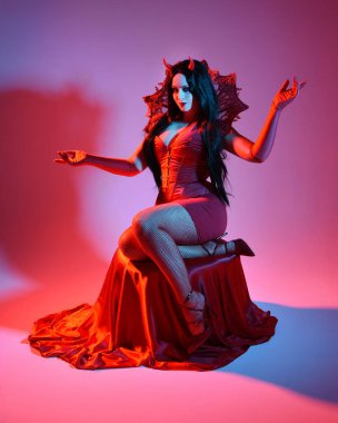 Gotik fantezi burlesk cadılar bayramı temalı şeytan kostümlü kırmızı elbise giyen güzel bir kadın manken portresi, boynuz ve eldiven. izole oturma pozisyonu, dramatik renkli stüdyo ışıkları