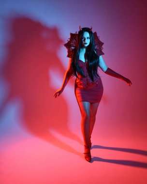 Gotik fantezi burlesk cadılar bayramı temalı şeytan kostümü, boynuz ve eldivenli kırmızı elbise giyen güzel kadın mankenin tam boy portresi. Zarif ayakta duruş, dramatik renkli stüdyo ışıkları.