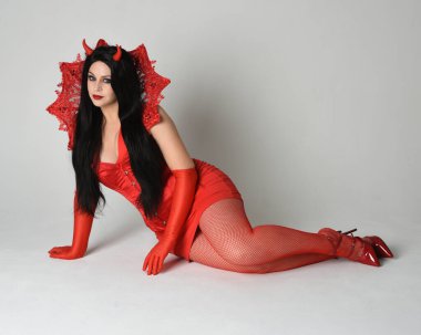 Gotik fantezi burlesk cadılar bayramı temalı şeytan kostümlü kırmızı elbise giyen güzel bir kadın manken portresi, boynuz ve eldiven. izole oturma pozisyonu, dramatik renkli stüdyo ışıkları