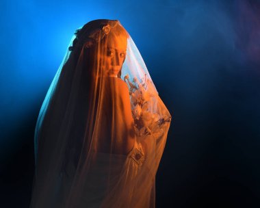 Güzel bir kadın modelin, ruhani bir başlık takan, duvak takan, ürkütücü bir hayalet gelin kostümü giyen sinematik portresi. Esrarengiz duman efektleriyle, elinde çiçeklerle karanlık stüdyo arka planı.